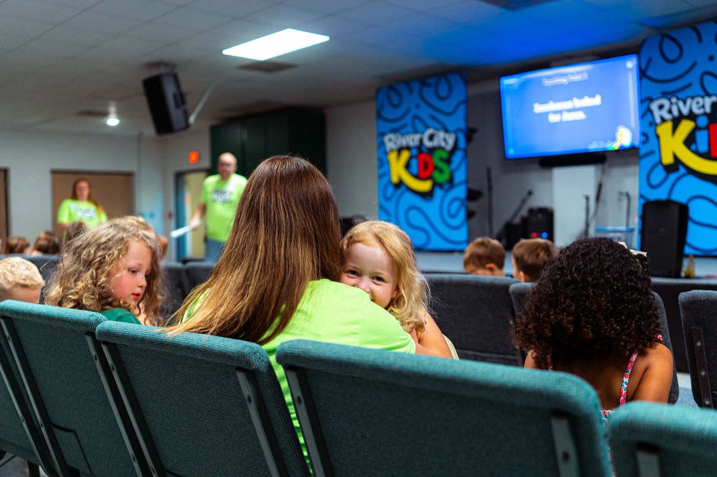 A leader holding a small child in the kids room, others seated in front of the River City Kids screen